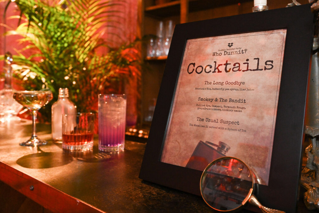 Cocktails sit on a table next to a sign that lists the murder-mystery-themed names of those cocktails