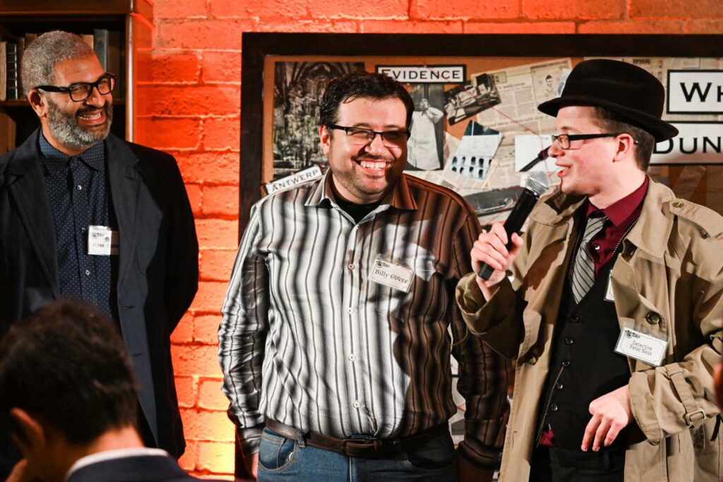 A smiling detective interviews two laughing men in a suspect lineup at a murder mystery party event.