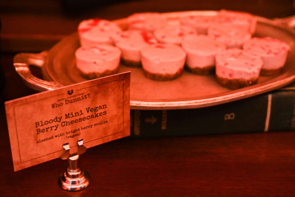 Tiny cheesecake bites sit on a plate next to a sign that gives them a clever, murder-mystery-themed name.